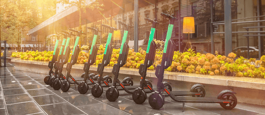 Row of electric scooters parked on a city sidewalk near a glass building and greenery in sunlight