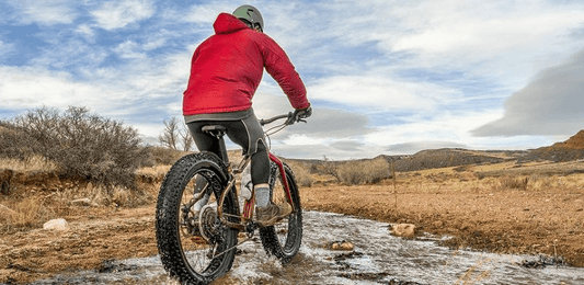 Person riding a red electric bicycle with fat tires through a shallow stream in an outdoor rugged landscape