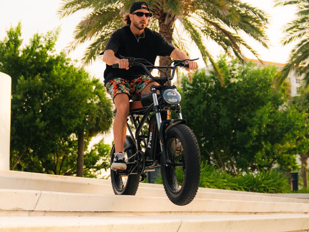 A man riding a hing-quality, black Freego fat-tire F2 Pro electric bike down the stairs, with the palm tree and other greenery in the background