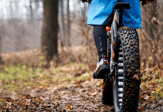 Close-up of a rider on a fat e-bike with large tires navigating a forest trail with fallen leaves and rough terrain.