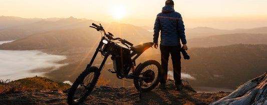 A man standing behind a Freego electric bike parked on the edge of a mountain slope, looking at the rising sun. Freego - for moments totally worth the price