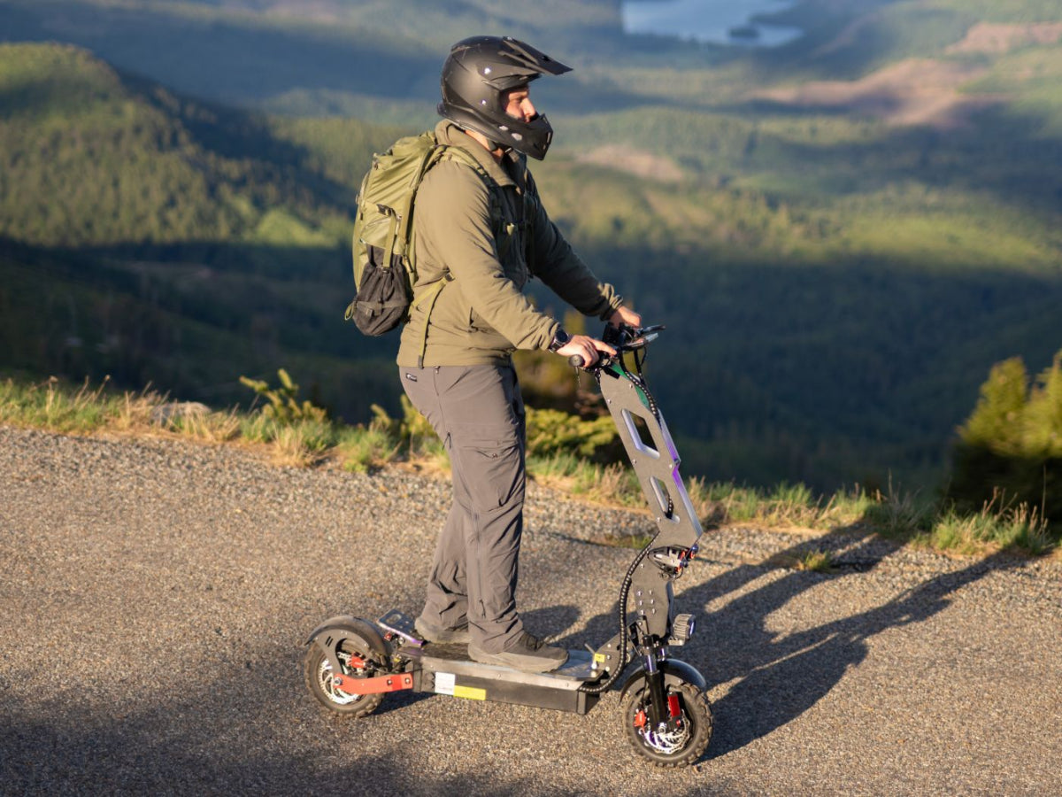 Person with a helmet and backpack standing on a mountain road with an electric scooter.