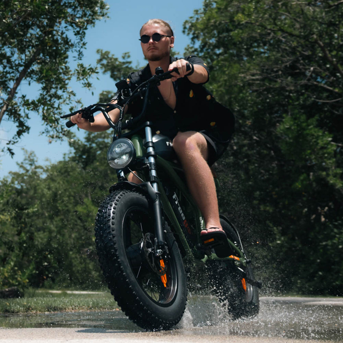 Person riding a green Freego F3 Pro electric bike through water with trees in the background