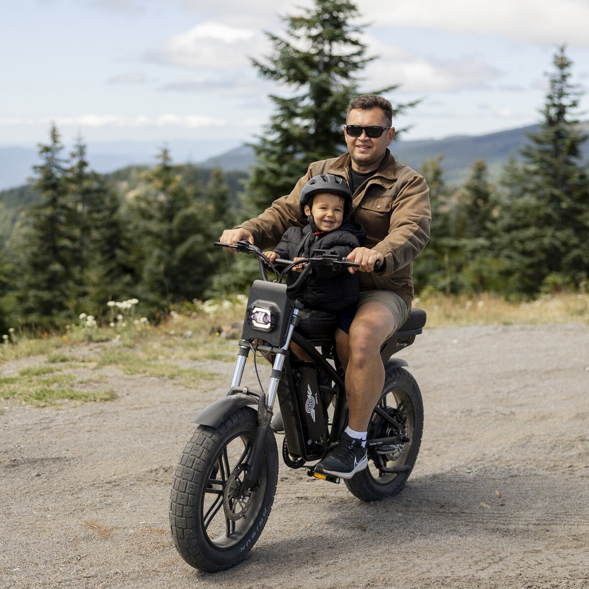 Man and child riding a small motorcycle on a road with trees and mountains in the background