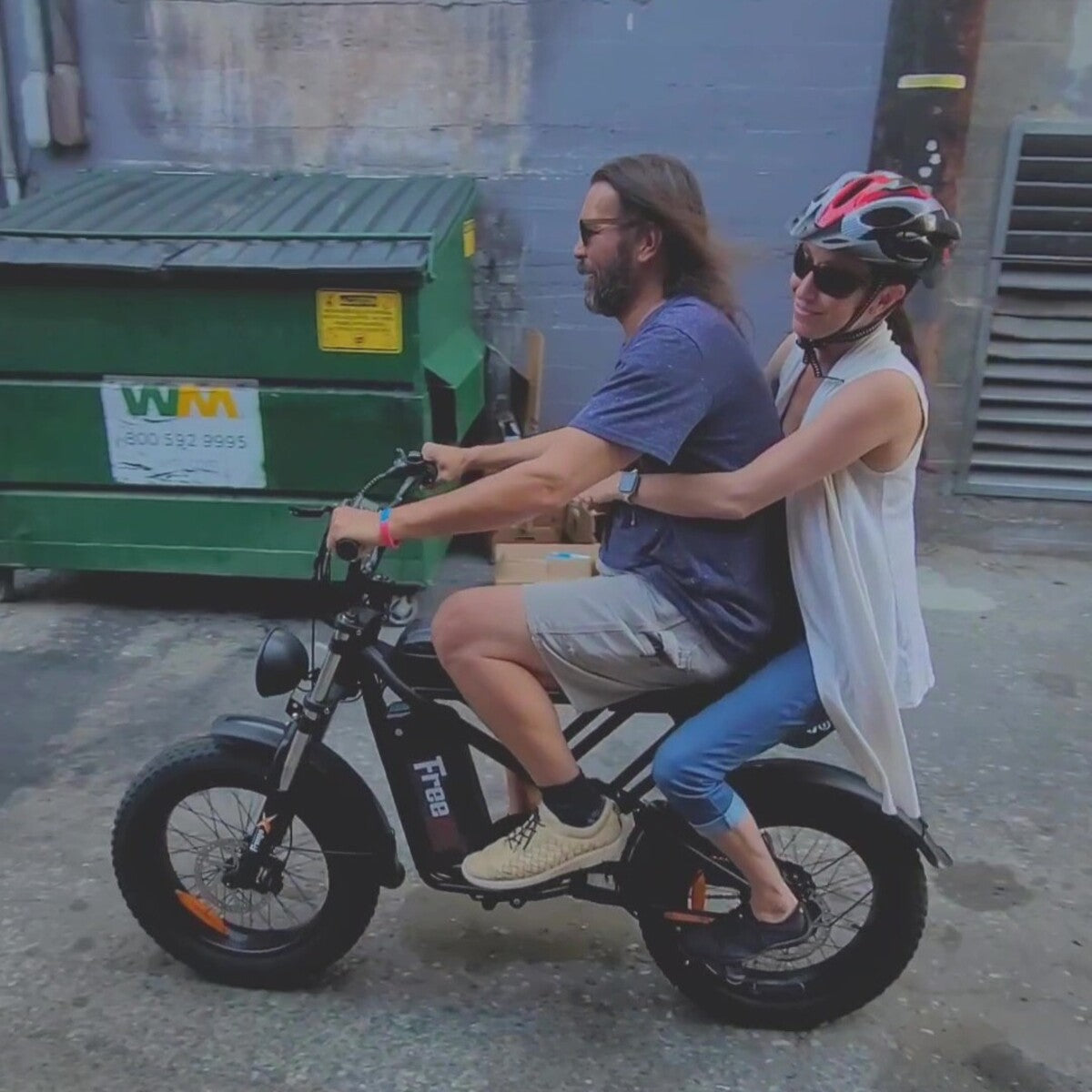 Two people riding an electric bike in an urban setting with a green dumpster in the background.