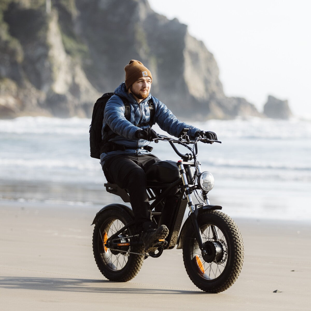 Person riding a Freego F3 Pro e-bike on a sandy beach with ocean and cliffs in the background