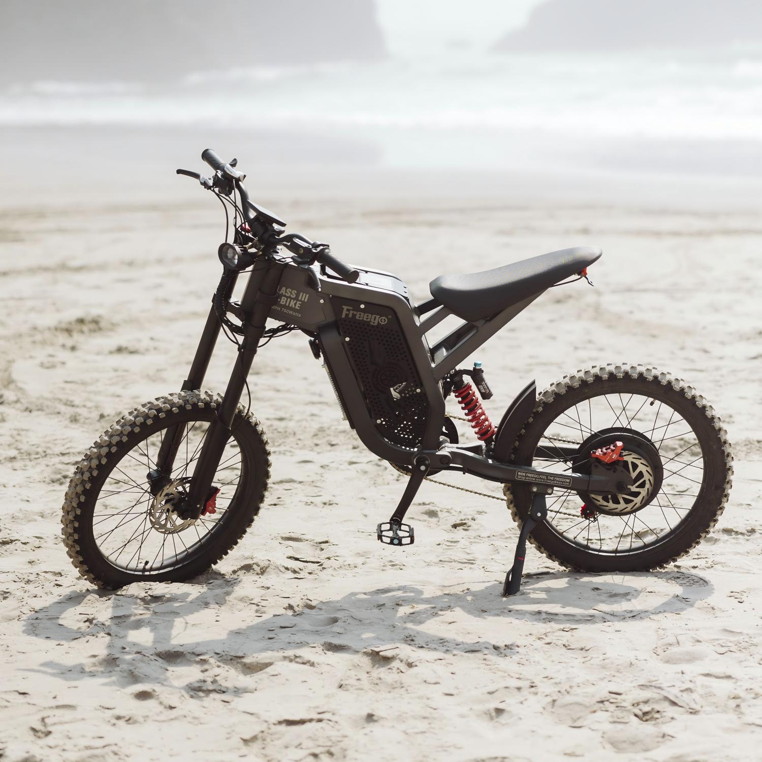 Freego electric dirt bike parked on a sandy beach, showcasing rugged off-road design against a scenic coastal backdrop.
