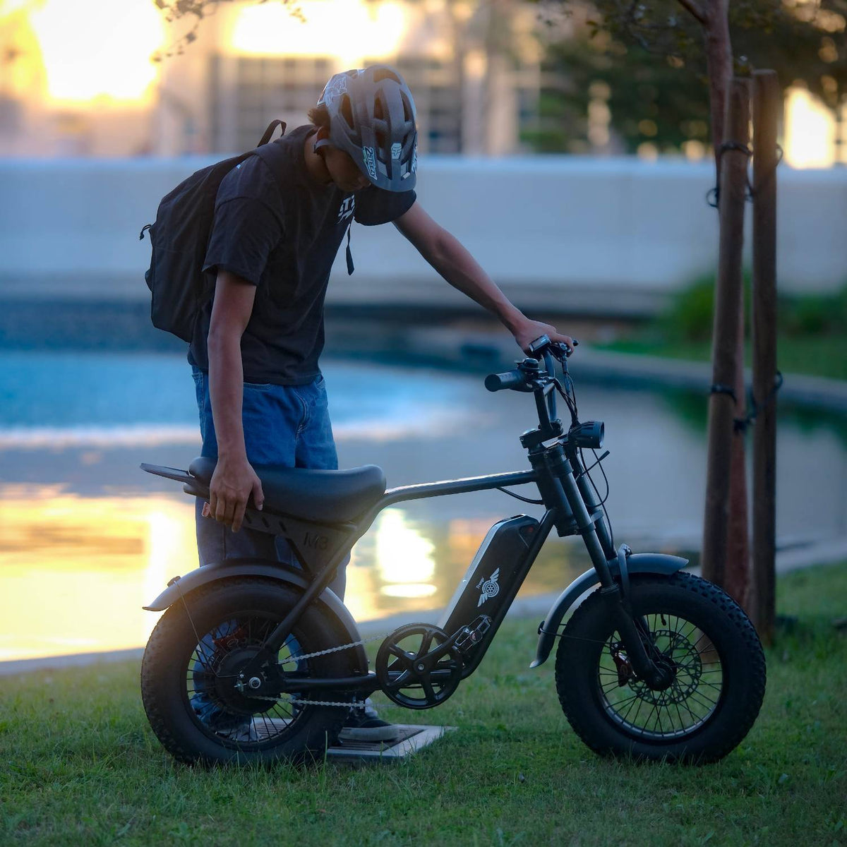 Helmeted rider standing over a Freego M3 electric mini bike by the water at sunset.