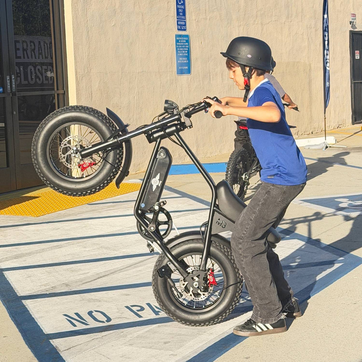 Helmeted rider lifting the front wheel of a Freego M3 electric mini bike while standing in a parking lot.