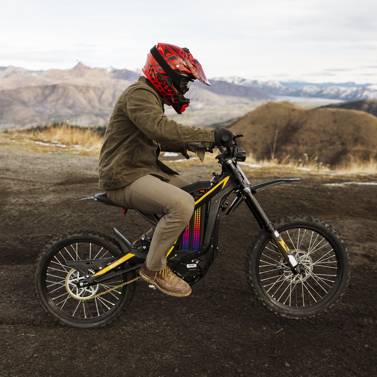 Side profile outdoor riding shot of rider with red helmet on Freego N1 electric dirt bike riding over mountain dirt terrain, rolling hills landscape background