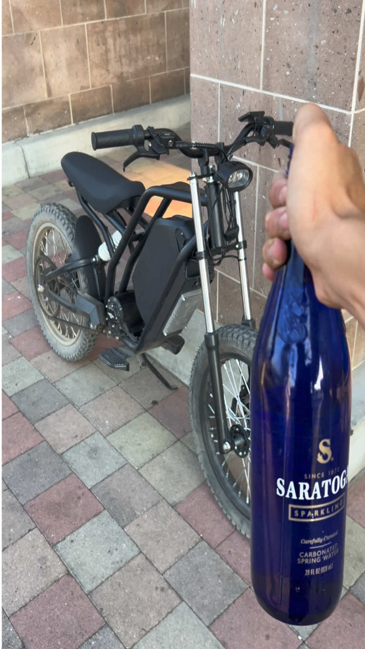 Person holding a blue bottle labeled 'Saratoga' next to a small motorcycle on a tiled patio.