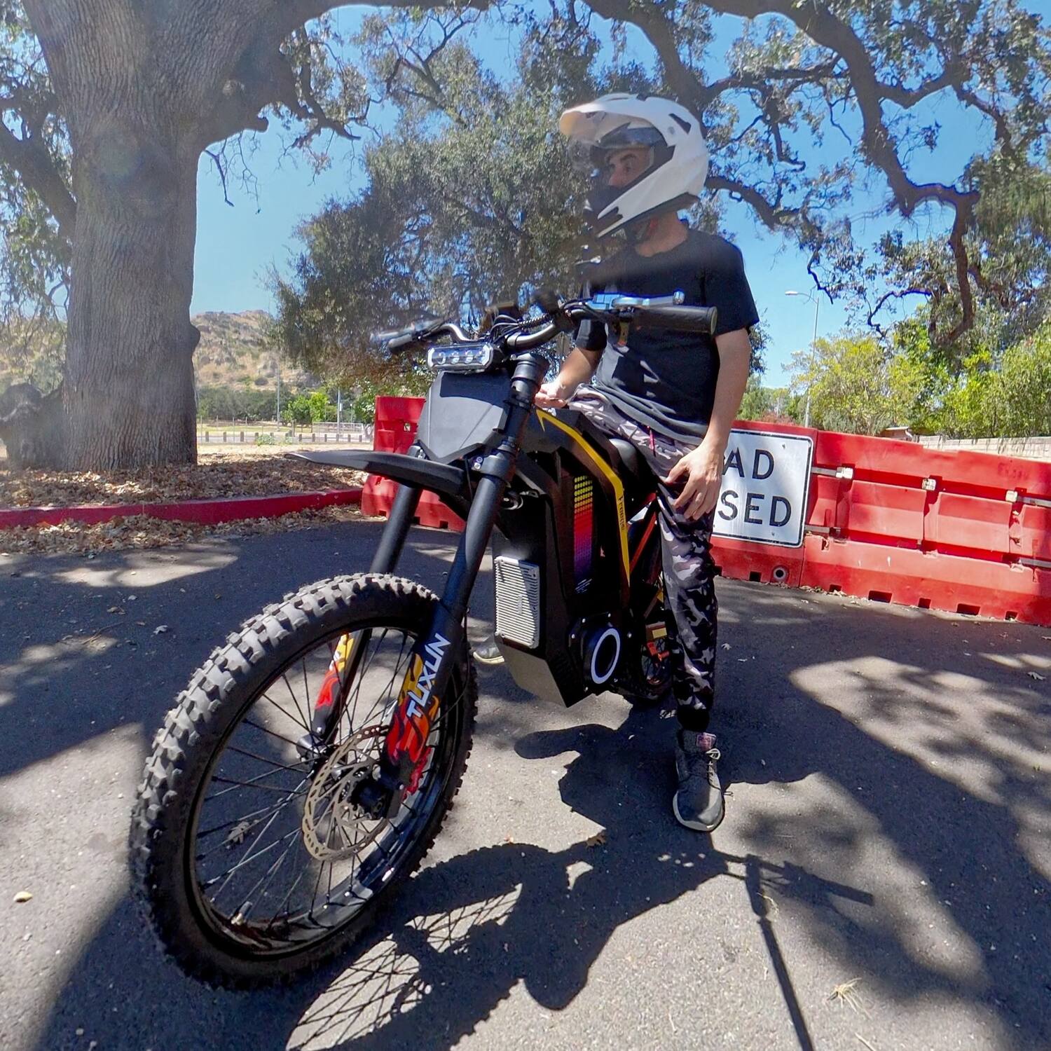 Lifestyle shot of rider with white helmet standing beside Freego Nova4 electric dirt bike under oak trees on sunny roadside