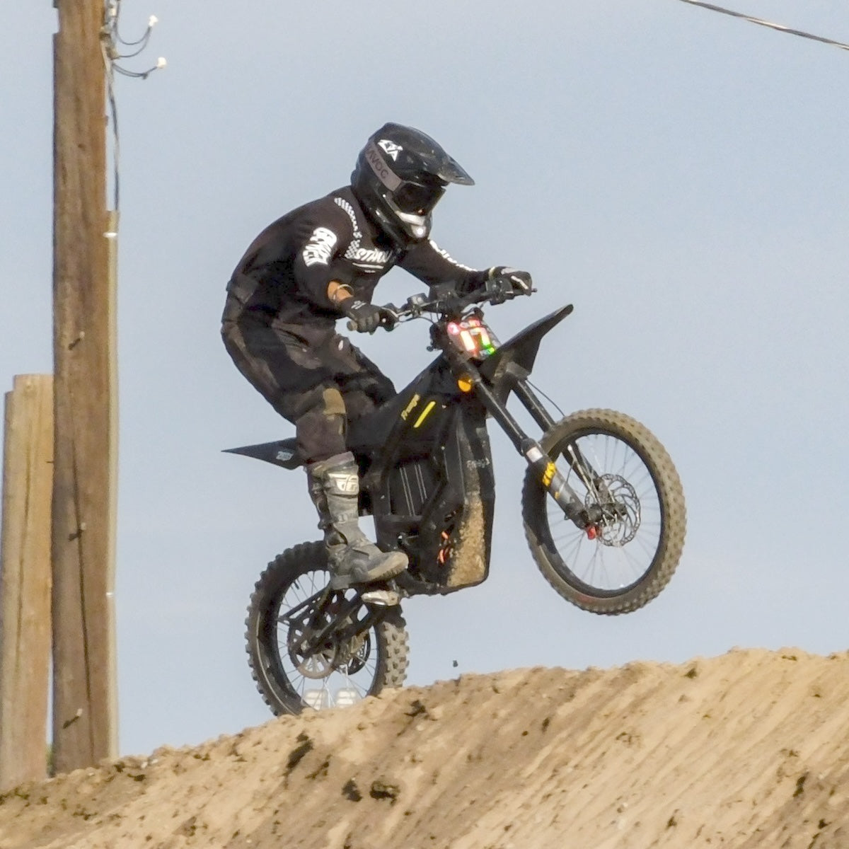 Rider in full black motocross gear jumping on Freego Nova5 electric dirt bike over sand track ramp, clear sky background