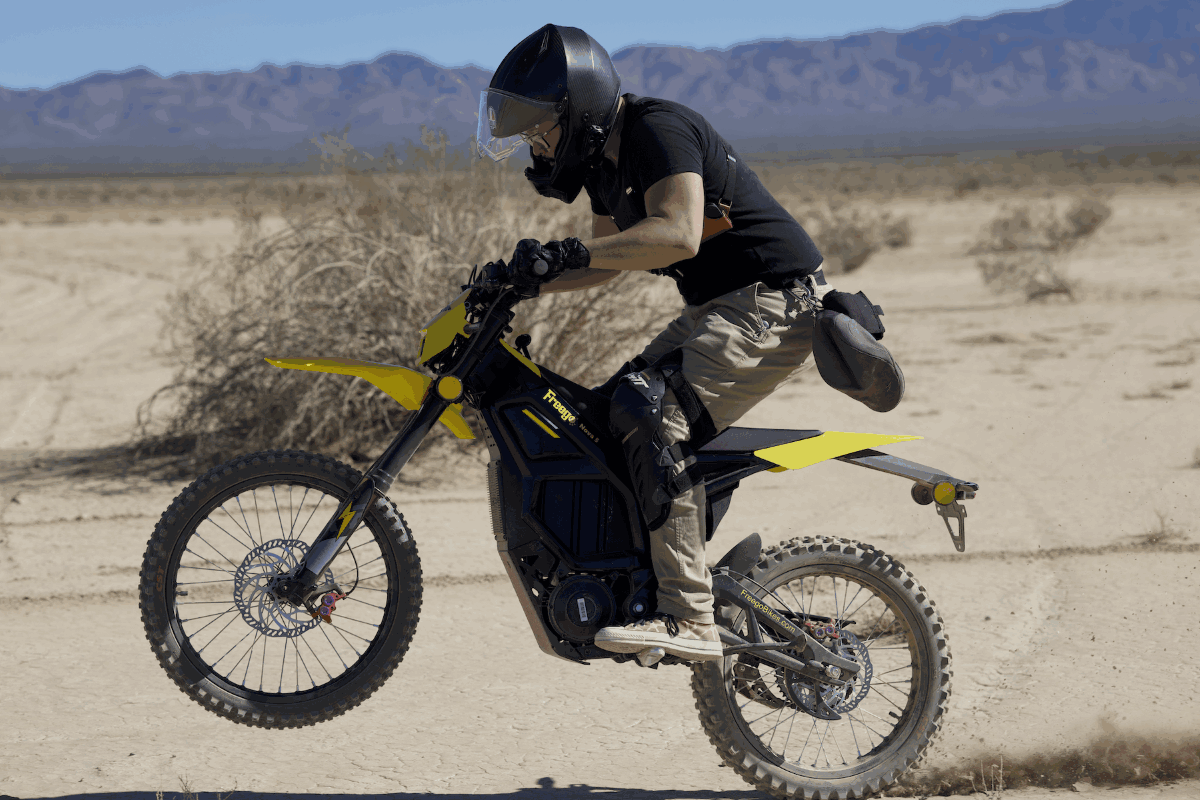 Person riding a black-and-yellow dirt bike Nova 5 in a desert landscape with mountains in the background