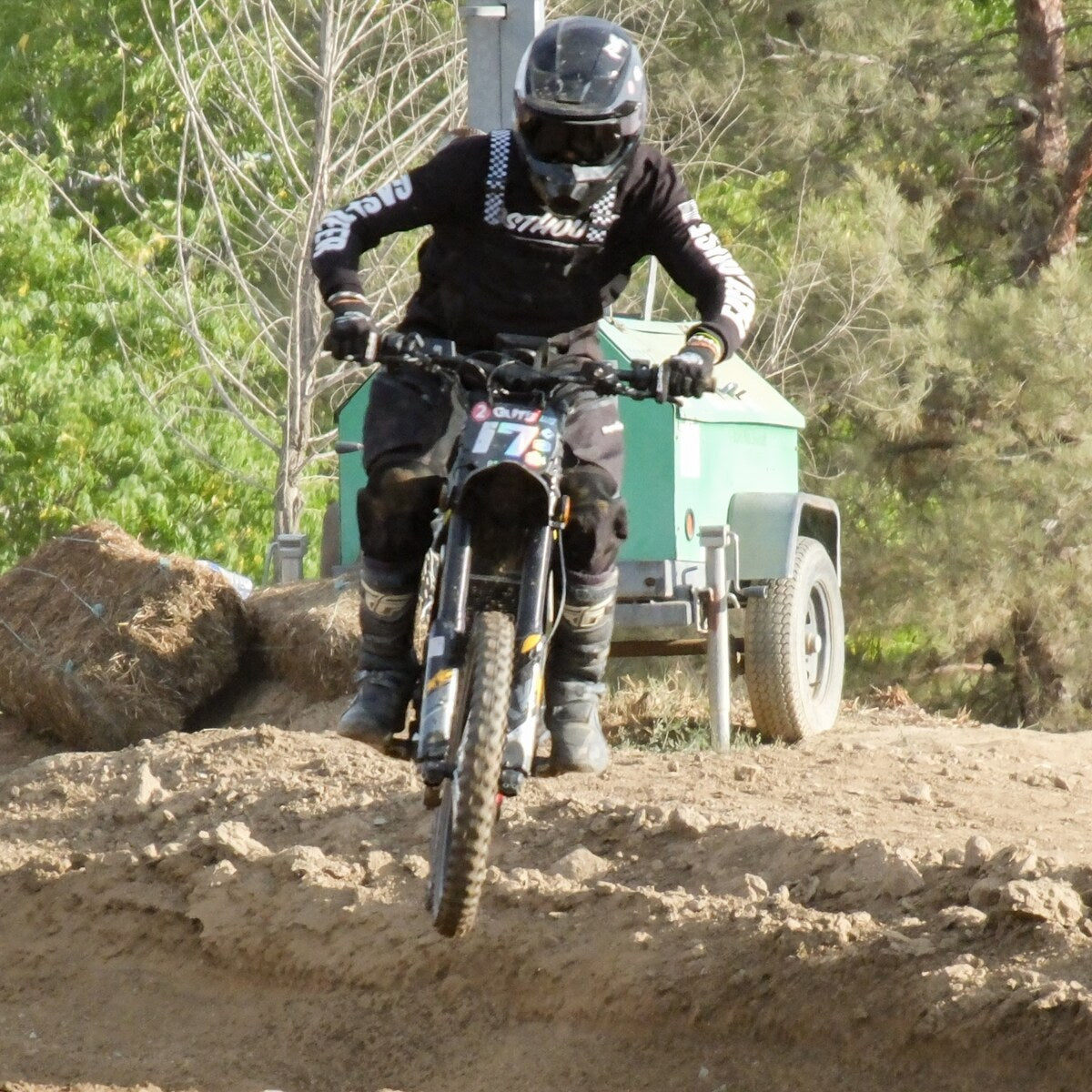 Front view of rider in full motocross gear jumping Freego Nova5 electric dirt bike over muddy race track, trees and utility trailer background.