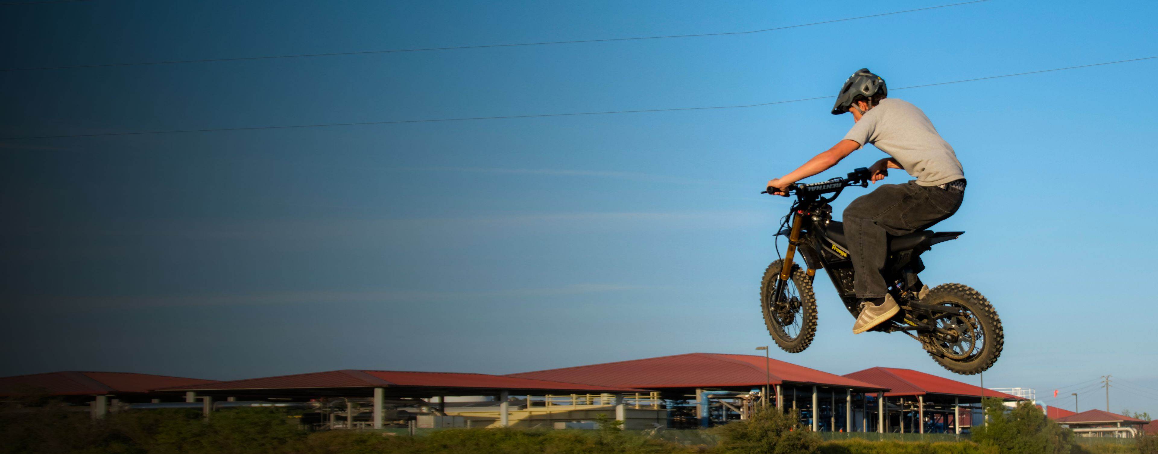 Person riding a Nova 5 Mini dirt bike in the air with buildings and clear sky in the background