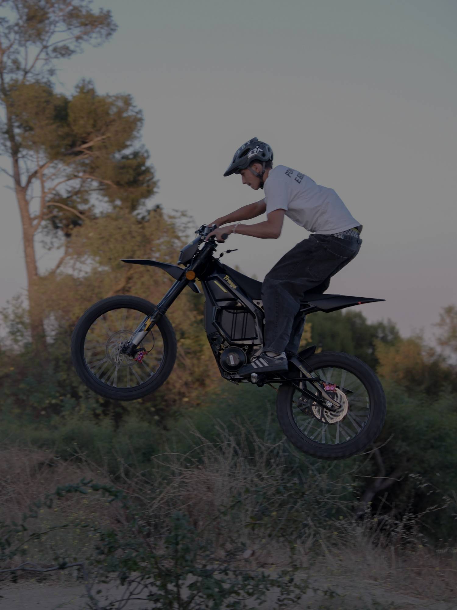 Vertical portrait shot of rider mid-air jump on Freego Nova5 electric dirt bike, dry hillside and trees background at soft dusk light.