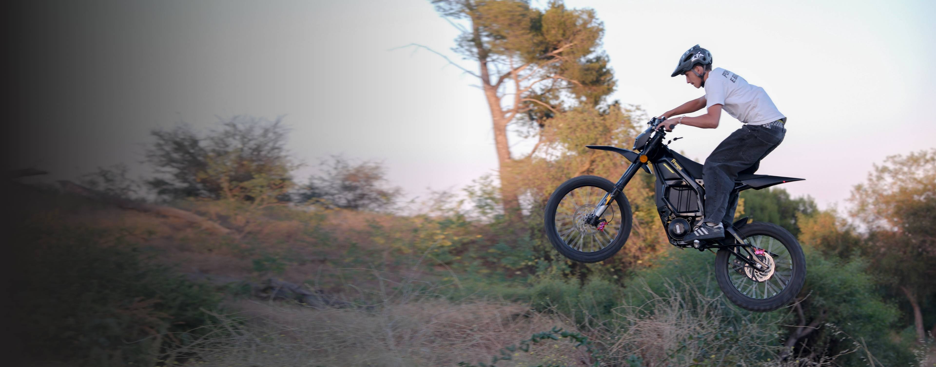 Panoramic banner shot of rider airborne jumping on Freego Nova5 electric dirt bike amid dry woodland hills at dusk, wide horizontal commercial banner image.