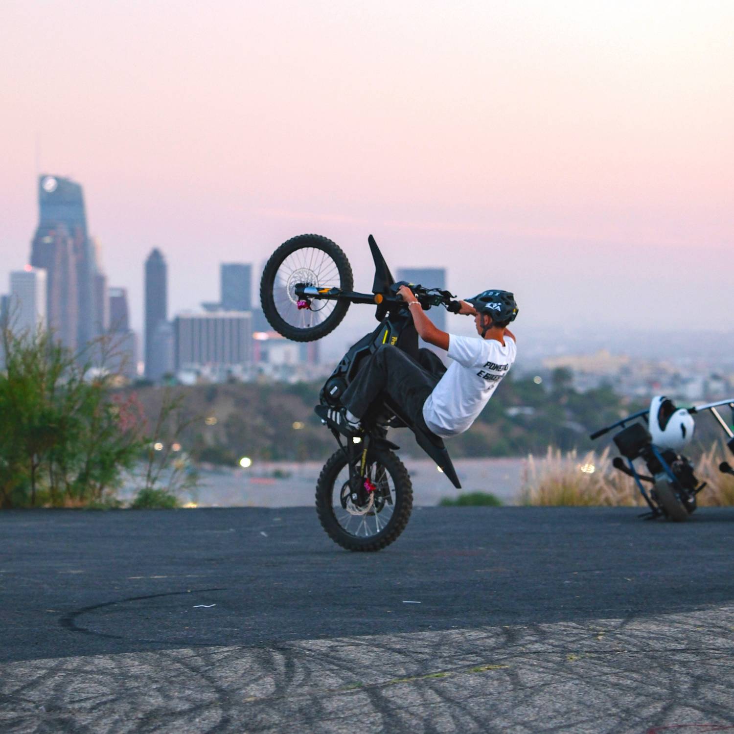 Rider doing wheelie on Freego Nova5 electric dirt bike, back view with city skyline at sunset