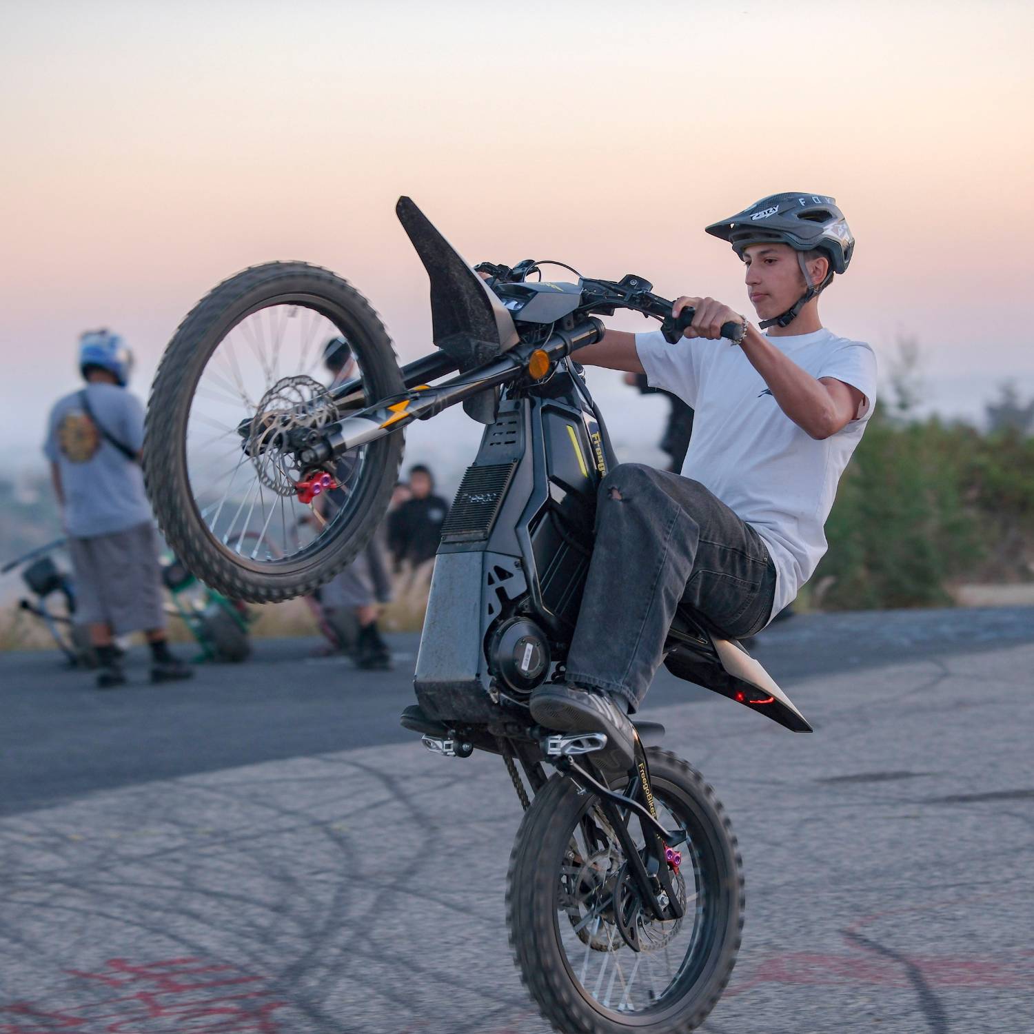 Close-up of rider performing wheelie stunt on Freego Nova5 electric dirt bike at sunset, with other riders in background