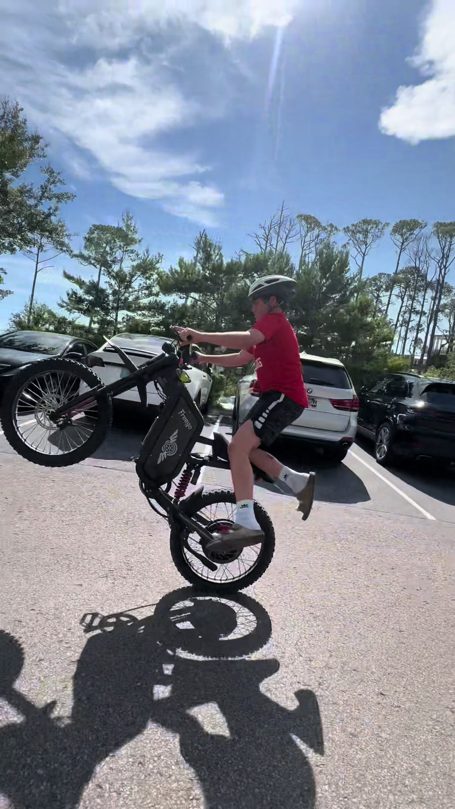 Vertical daytime action shot of rider doing wheelie stunt on black Freego electric bike in outdoor parking lot with pine trees and parked cars background