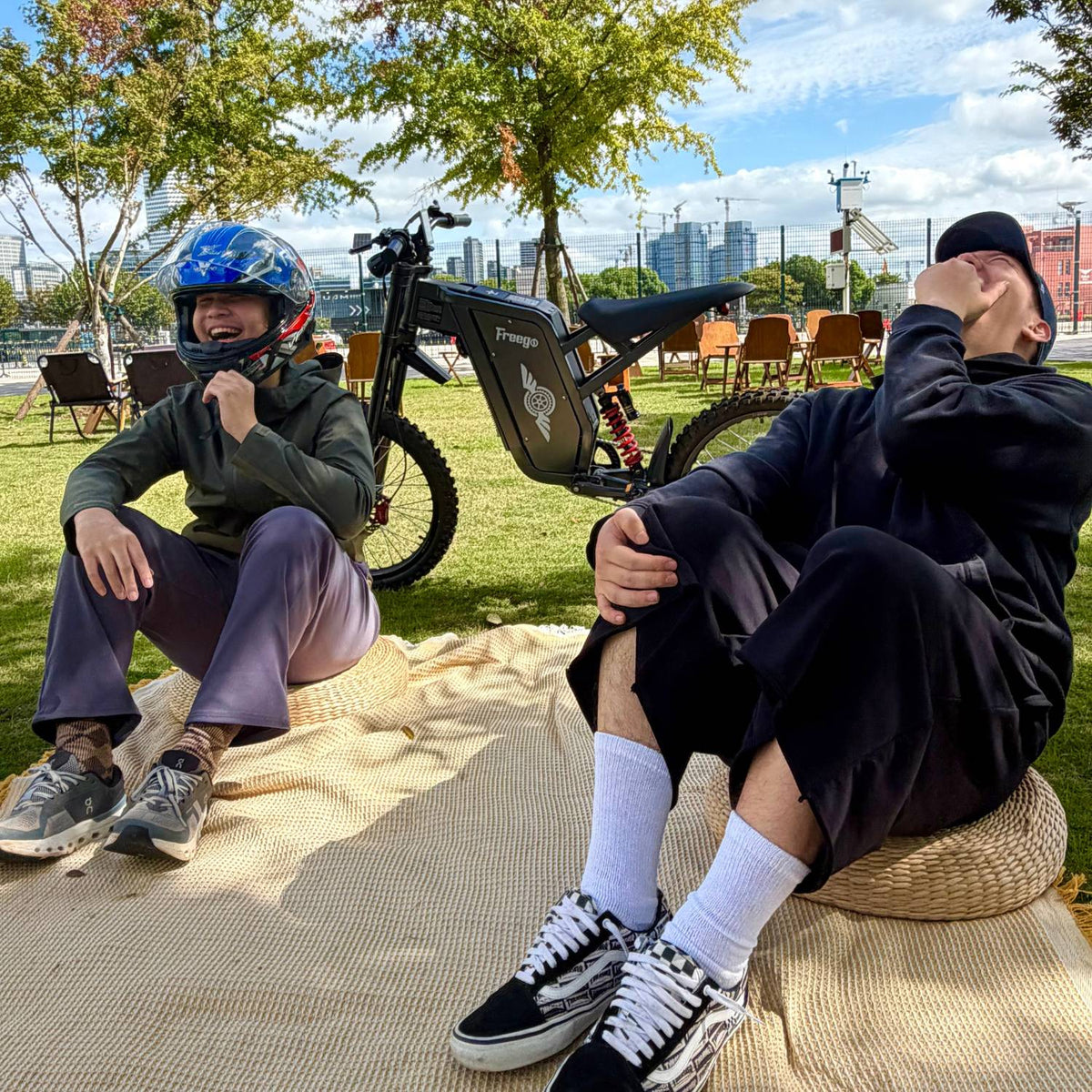 Two riders relaxing beside a Freego X2 Pro electric dirt bike in a sunny city park.