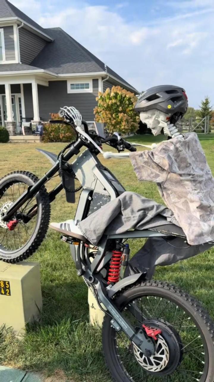 Skeleton sitting on a bicycle in a yard with a house in the background