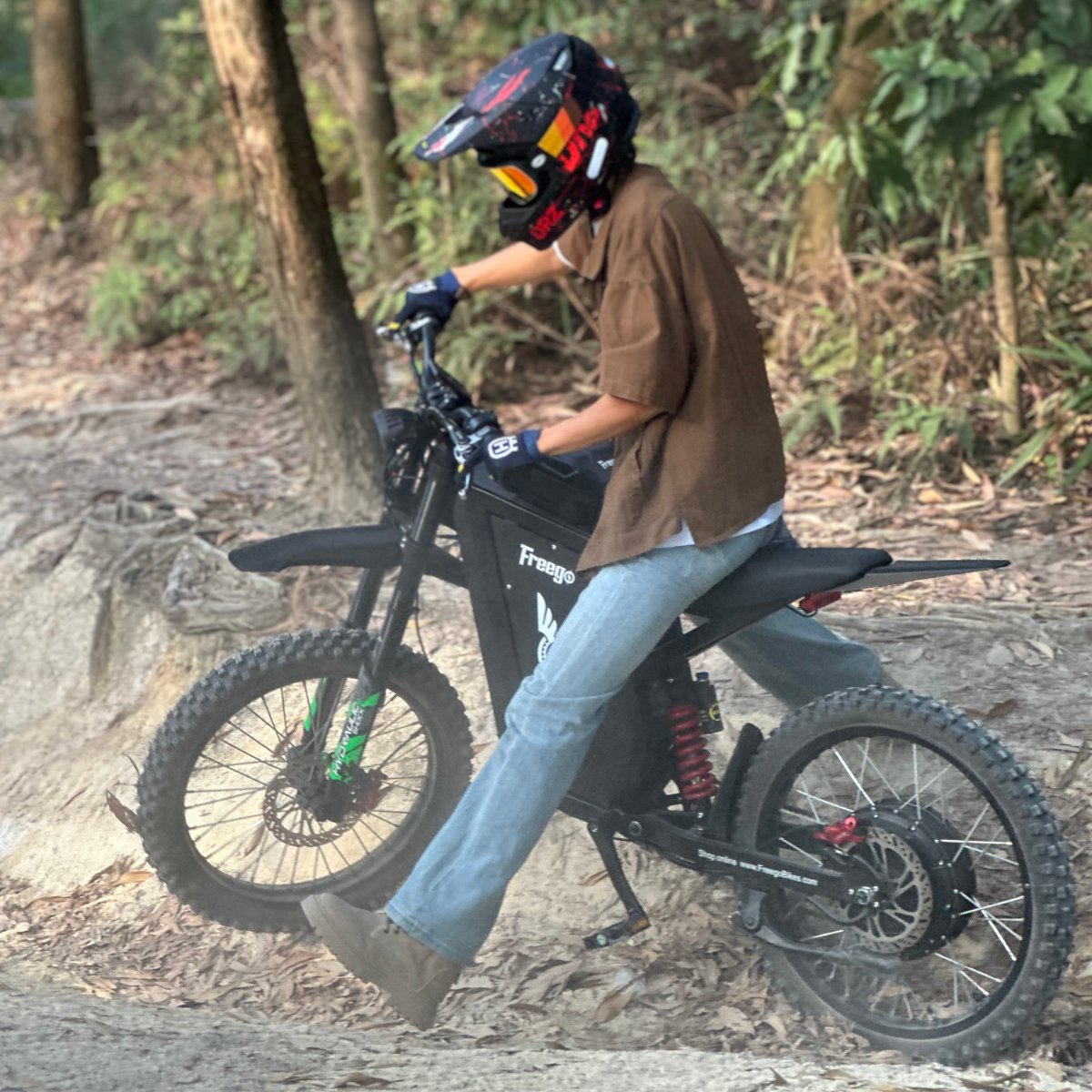 Freego X3 electric dirt bike (with pedals) leads the frame, riding a sunlit wooded trail; helmeted rider onboard, dust swirling under wheels.