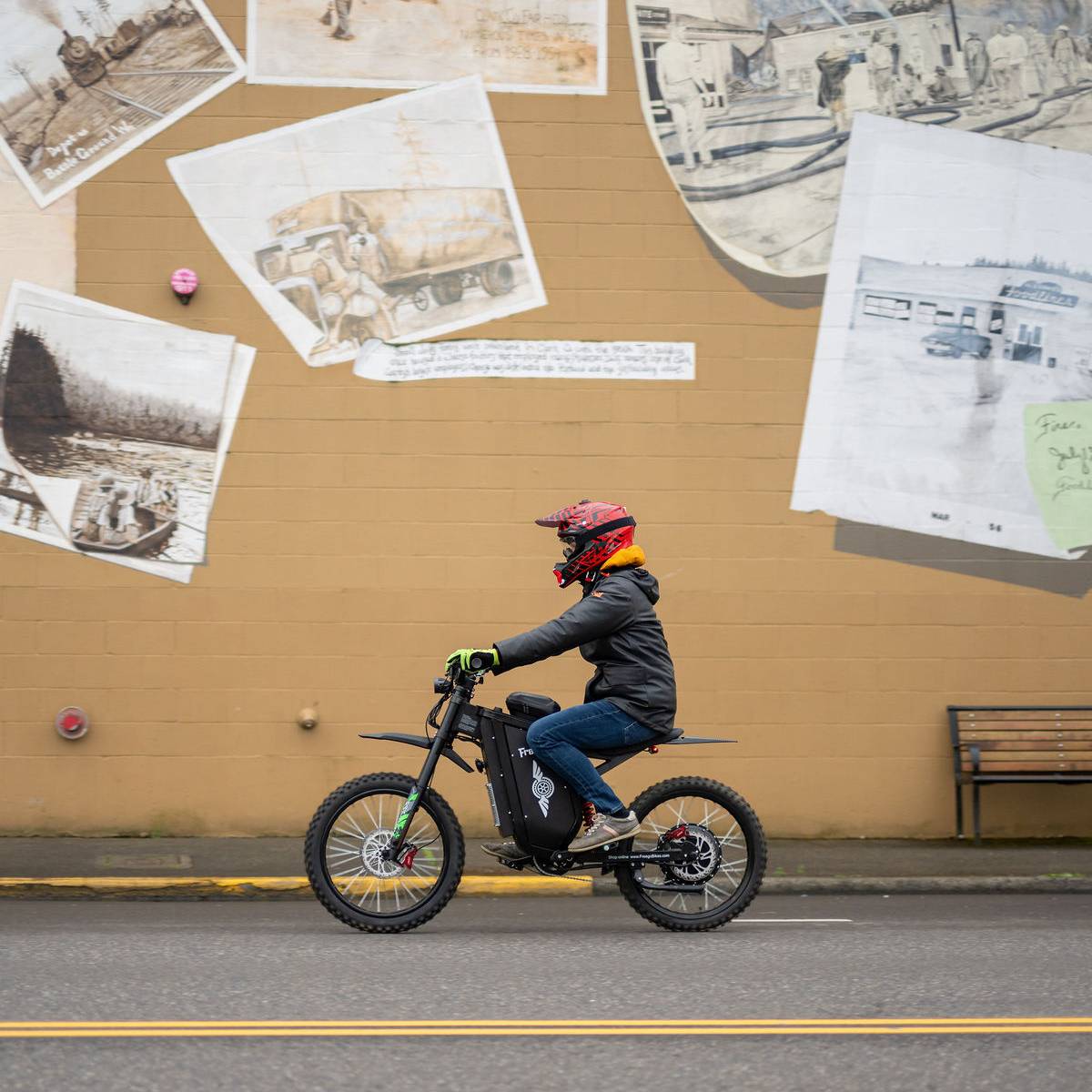 A person riding a black electric dirt bike on a street in front of a tan wall covered with vintage-style photographs and handwritten notes.