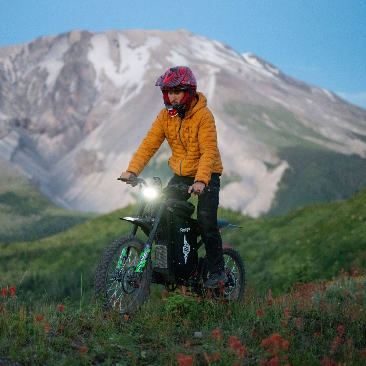 A person riding a black freego x3 electric dirt bike on a grassy mountain trail with a large snow-dusted mountain in the background. 