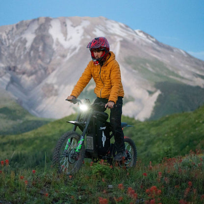 A person riding a black freego x3 electric dirt bike on a grassy mountain trail with a large snow-dusted mountain in the background. 