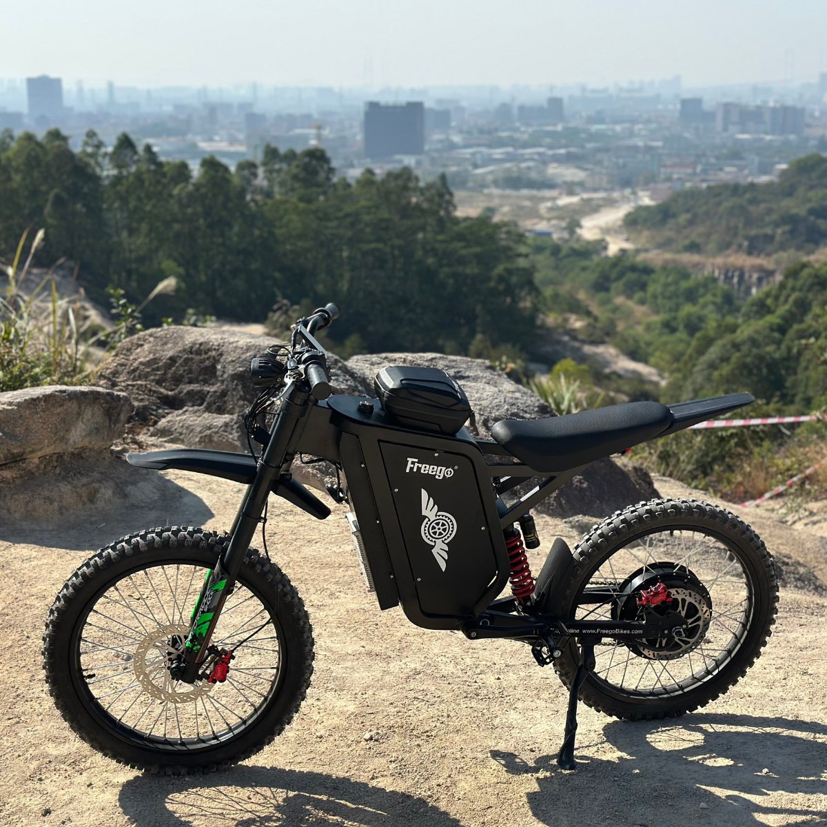 Freego X3 electric dirt bike (with pedals) parked on a rocky hill, city skyline and forested backdrop visible; brand logos and rugged tires on display.