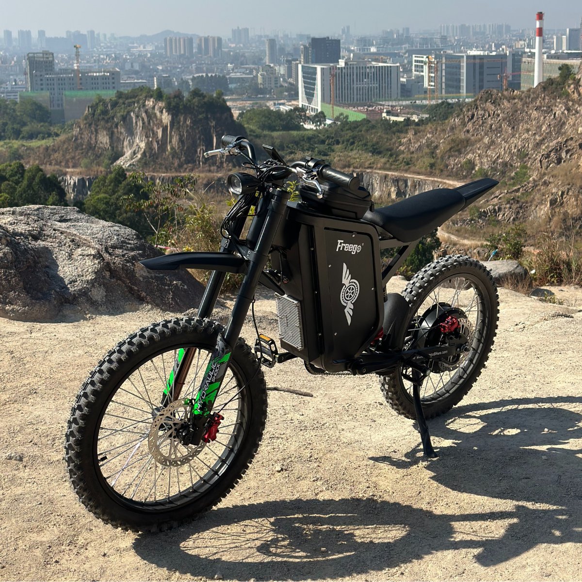 Freego X3 electric dirt bike is parked on a rocky hill at the city’s edge; its rugged tires and brand logo stand out, with urban buildings and quarried terrain in the backdrop.