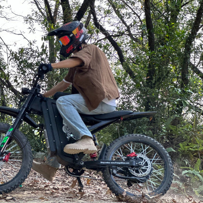 Freego X3 electric dirt bike (with pedals) navigates a leaf-strewn forest trail; helmeted rider controls the bike, surrounded by thick tree foliage.