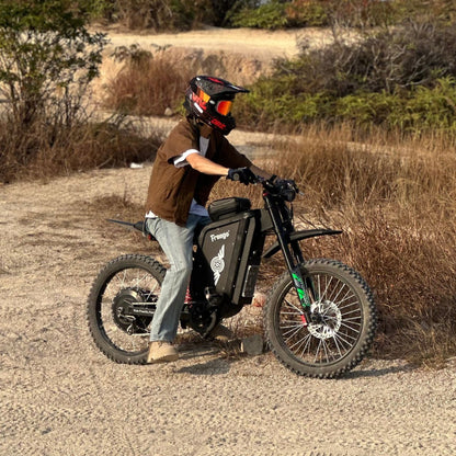 Freego X3 electric dirt bike (with pedals) is ridden on sandy ground, bordered by dry bush vegetation; helmeted rider grips the handlebars, bike’s brand logo visible.