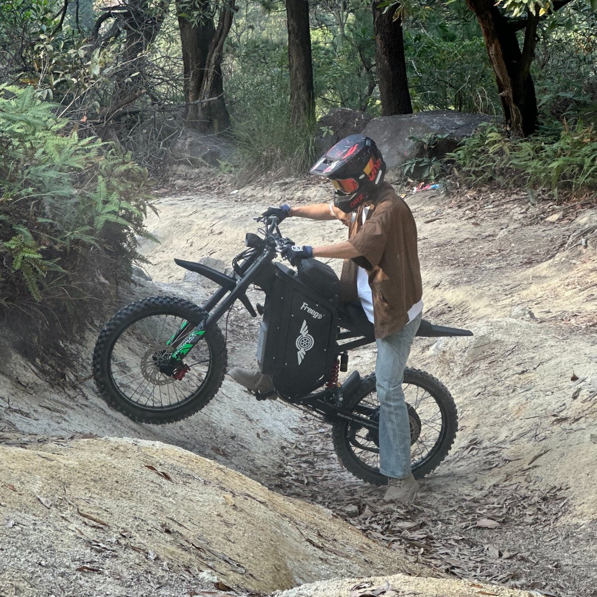 Freego X3 electric dirt bike navigates a rocky gully in a forested area; helmeted rider balances the bike, with ferns and trees lining the uneven trail.