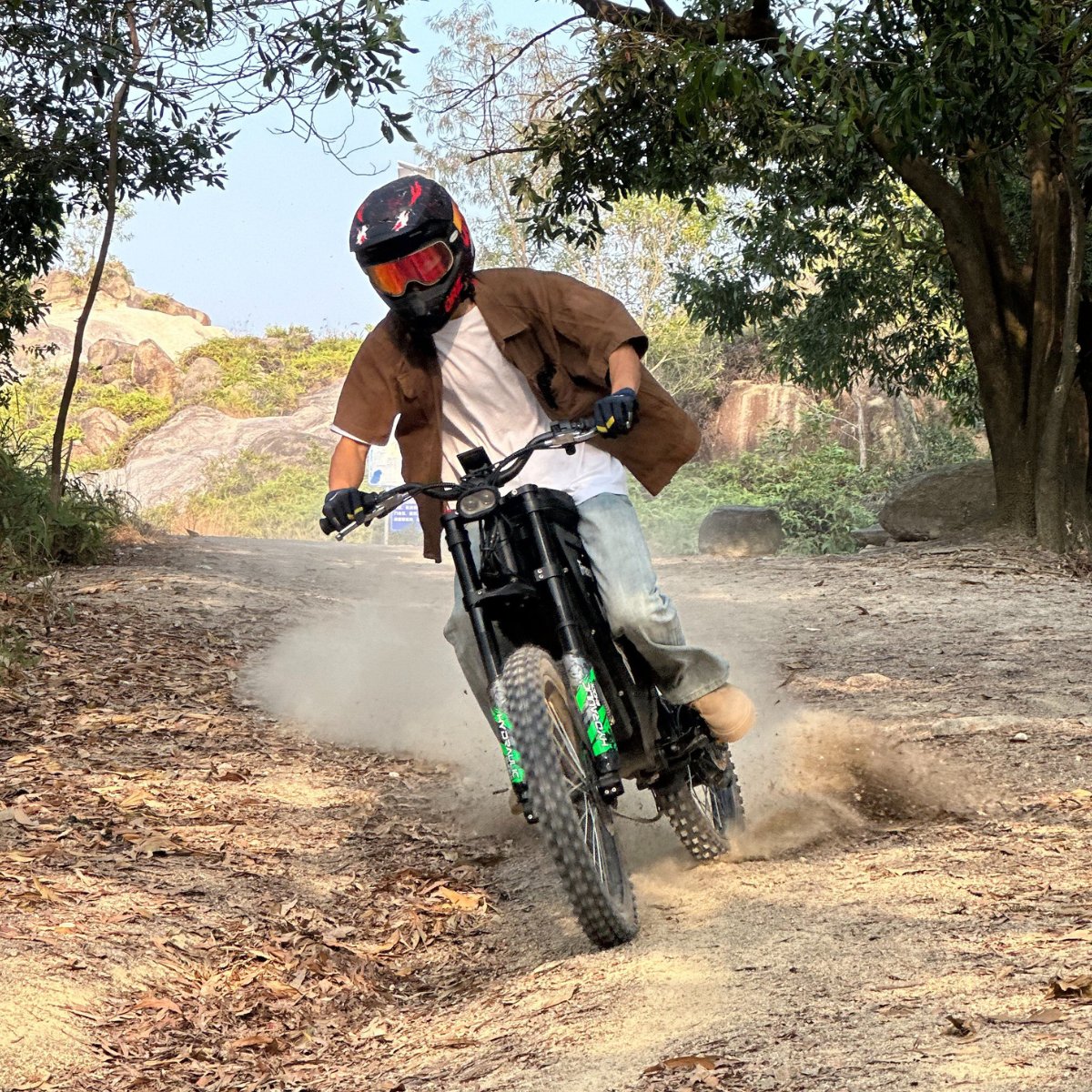 Freego X3 electric dirt bike (with pedals) kicks up dust on a leaf-littered, forested rock trail; helmeted rider steers the bike, with trees and boulders in the backdrop.