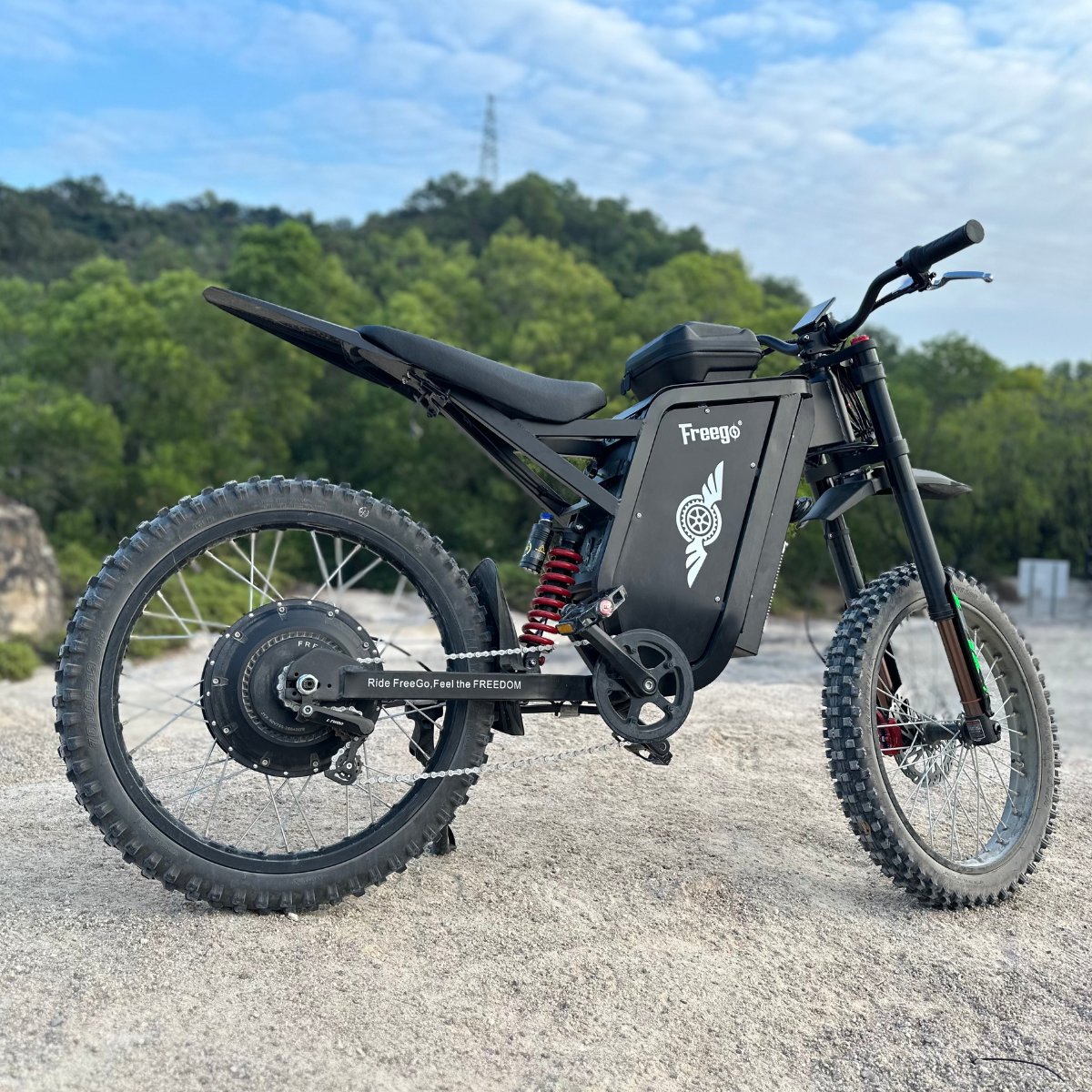 Freego X3 electric dirt bike rests on rocky ground, flanked by lush forest and a utility tower under a partly cloudy sky; its brand logos and rugged tires are prominent.