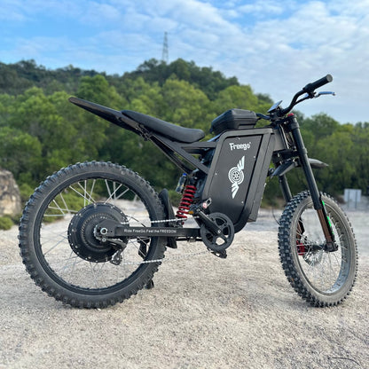 Freego X3 electric dirt bike rests on rocky ground, flanked by lush forest and a utility tower under a partly cloudy sky; its brand logos and rugged tires are prominent.
