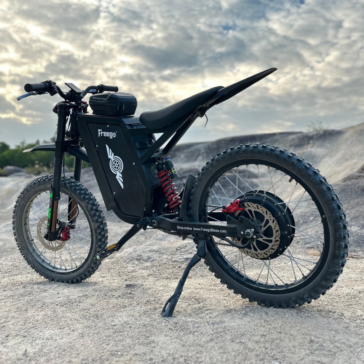 Freego X3 electric dirt bike (with pedals) parked on rocky terrain, dramatic sky backdrop; brand logos and rugged tires visible.