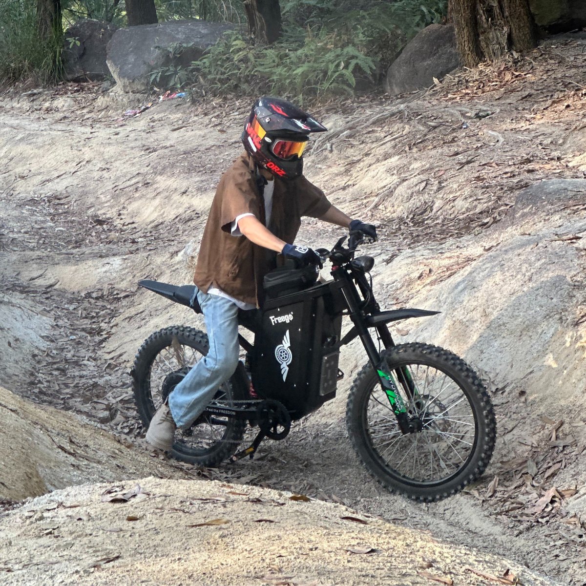 Freego X3 electric dirt bike (with pedals) climbs a rocky-sloped trail; helmeted rider maneuvers the bike, with forested rock terrain in the background.