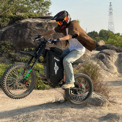 Freego X3 electric dirt bike (with pedals) lifts off a rock on rocky terrain; helmeted rider controls the bike, utility tower in the distance.
