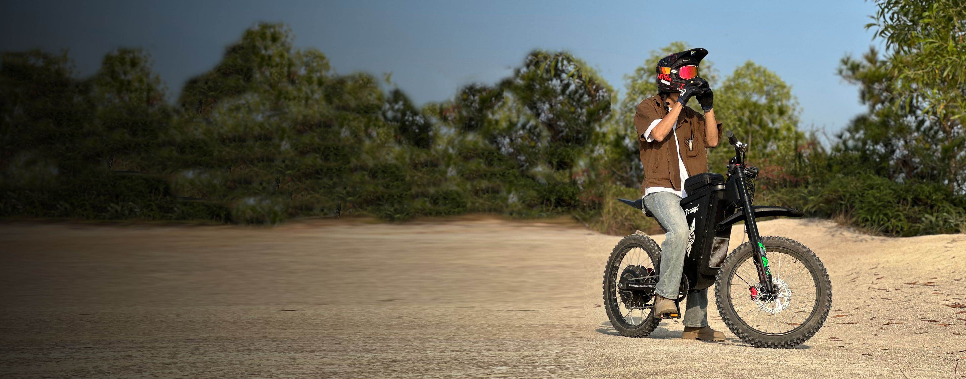 Freego X3 electric dirt bike is positioned on sandy terrain; a helmeted rider (in brown shirt) operates filming equipment beside it, with dense greenery and clear sky in the backdrop.