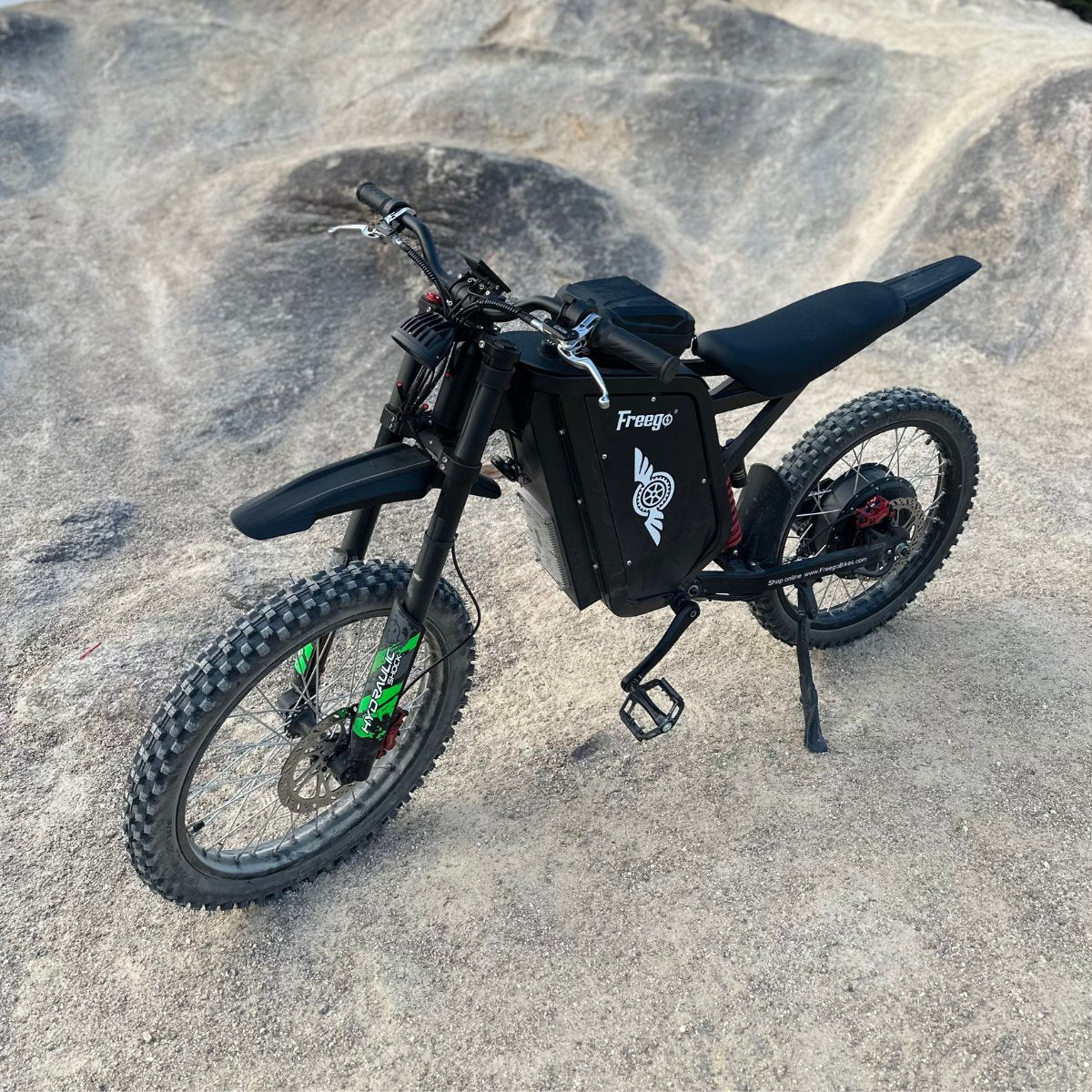 Freego X3 electric dirt bike (with pedals) rests on smooth rocky terrain; its rugged tires, brand logo, and pedal system are clearly visible.