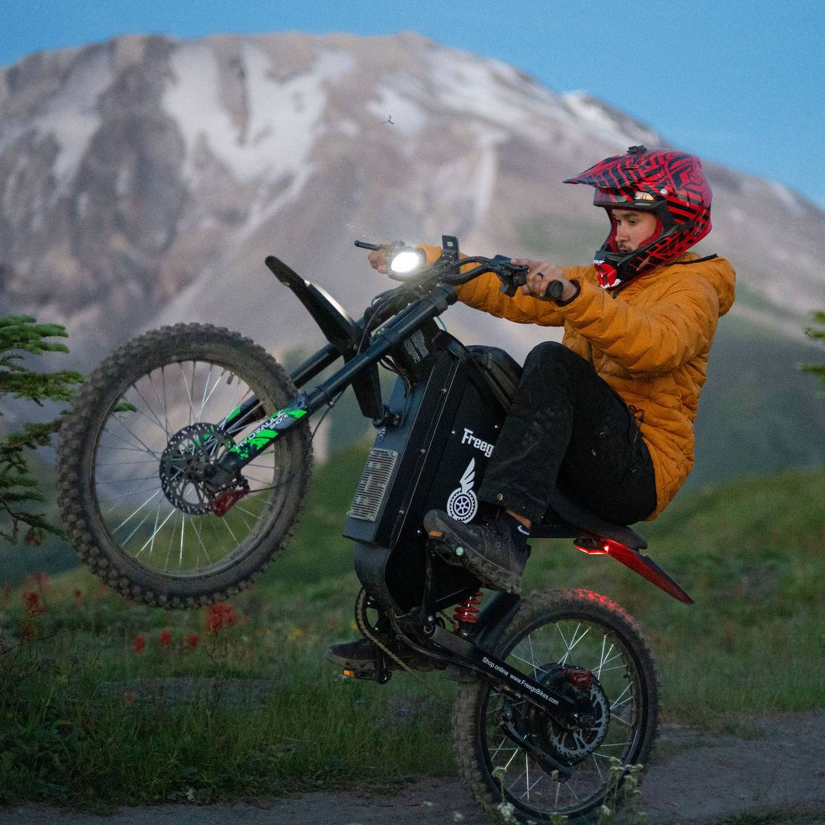 A person wearing a red and black helmet and a yellow jacket rides a black freego x3 electric dirt bike on a mountain path with a snowy mountain in the background.
