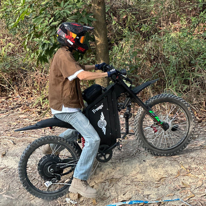 Freego X3 electric dirt bike ascends a leaf-covered wooded slope; helmeted rider maneuvers the bike, with dense undergrowth in the background.