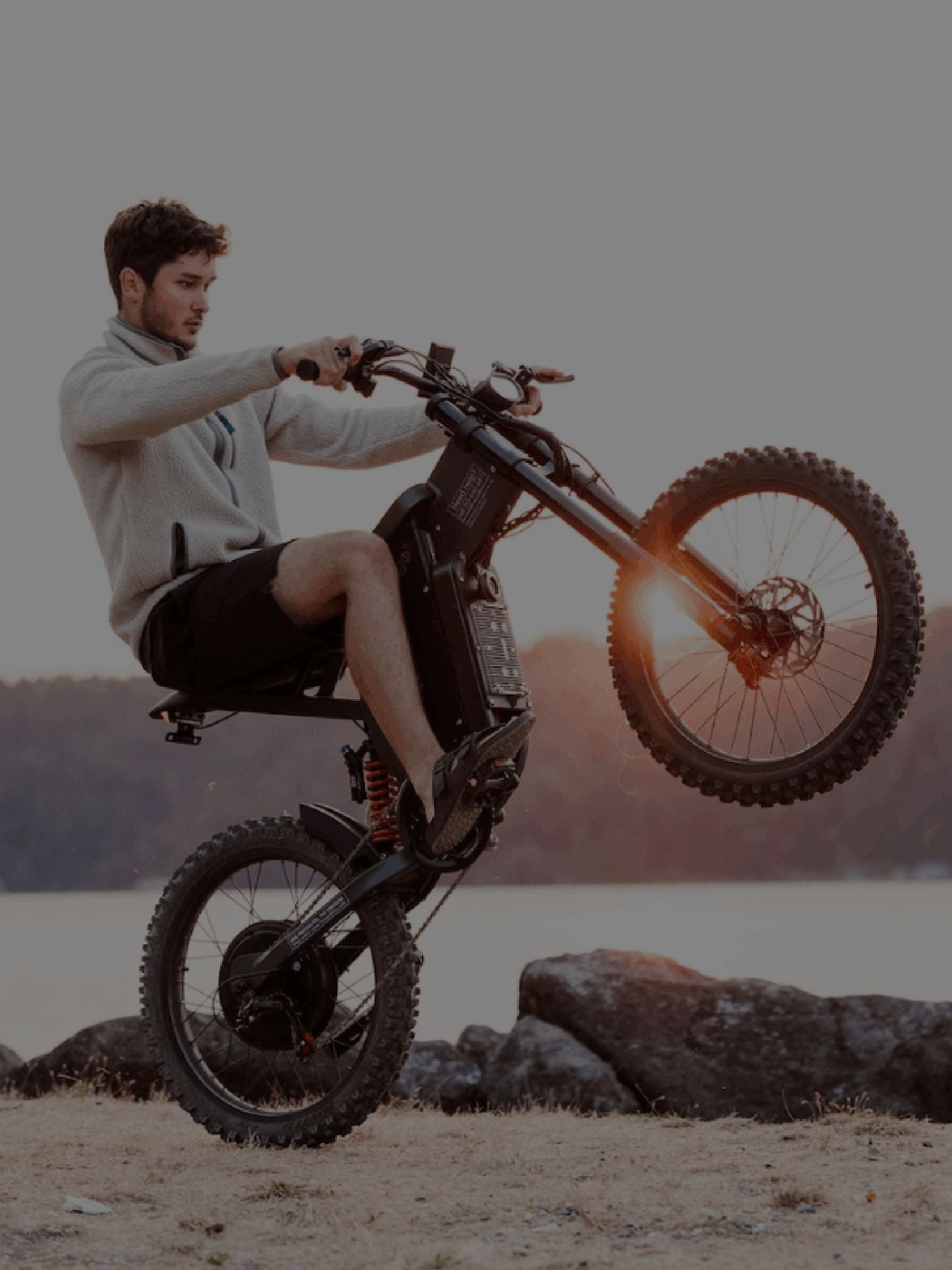 Man dressed in a jacket and shorts doing wheelie by shore.