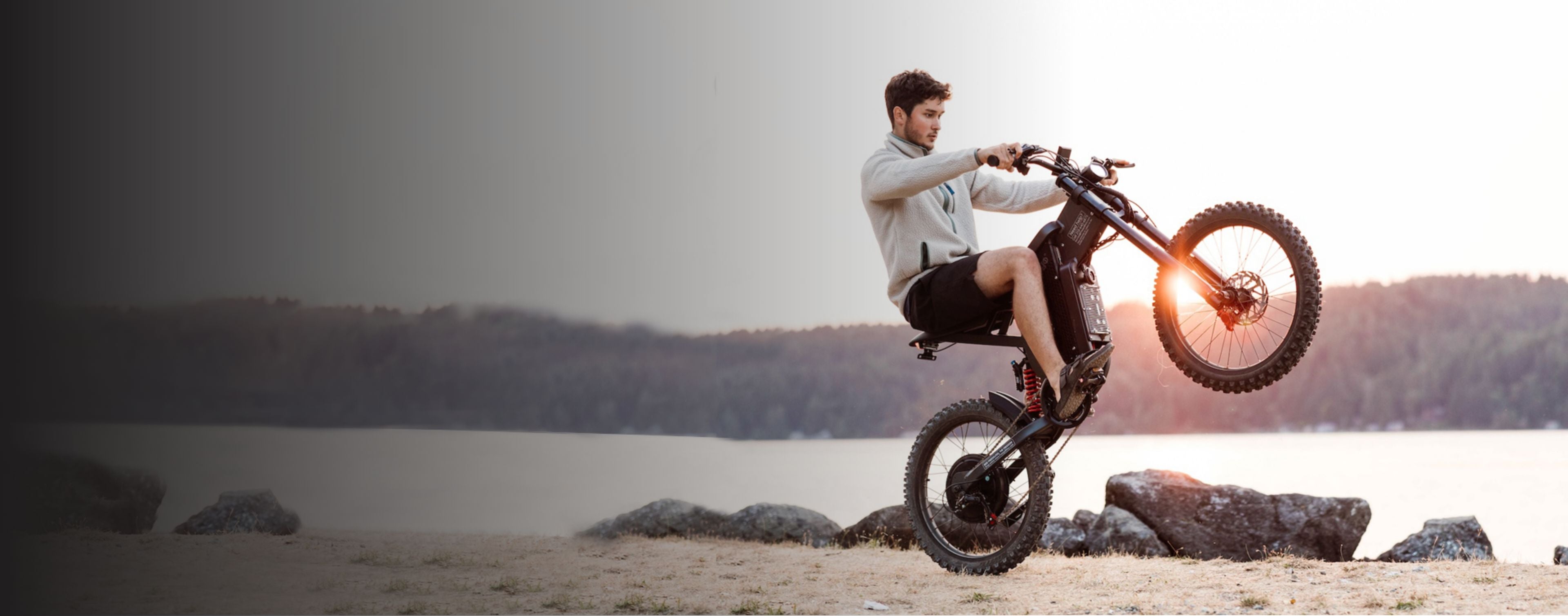 A man wearing a jacket and shorts performs a wheelie on a Freego X2 on flat ground near the shoreline, with dramatic lighting effects.