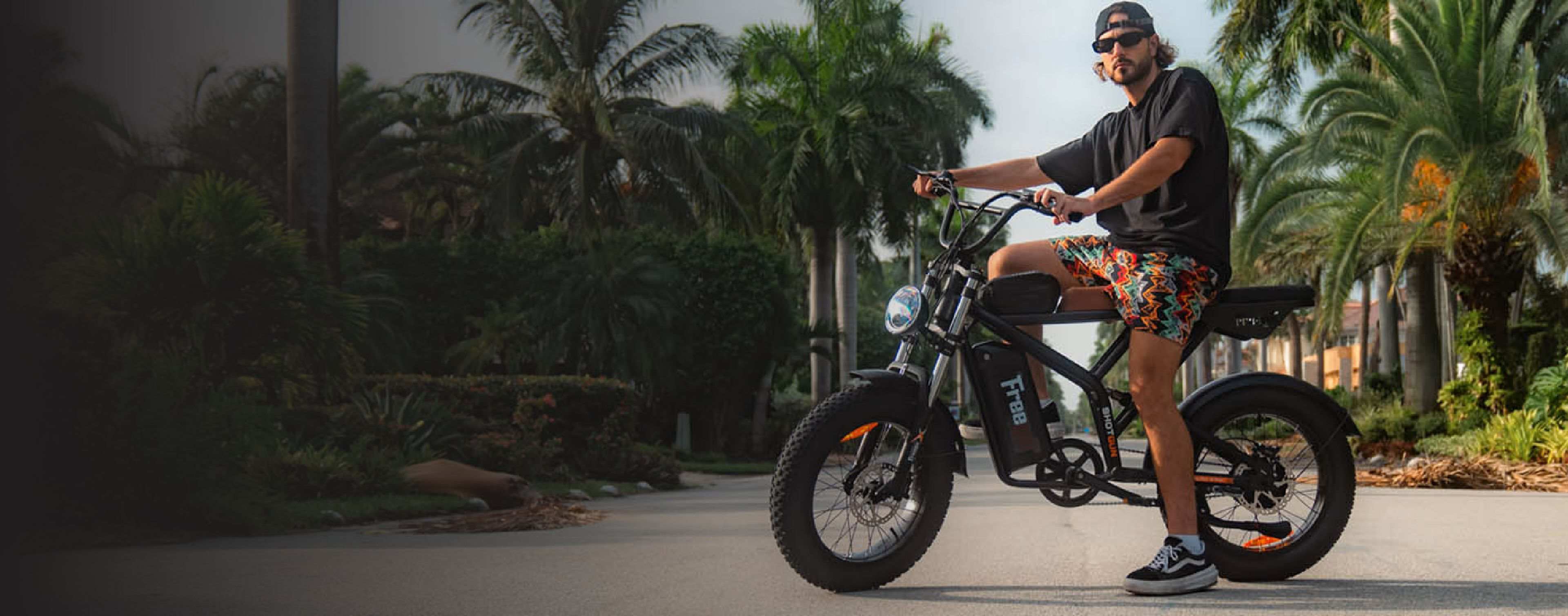 A man wearing floral pants, sunglasses, and a baseball cap poses with the F2 Pro e-bike in the center of a palm-lined road