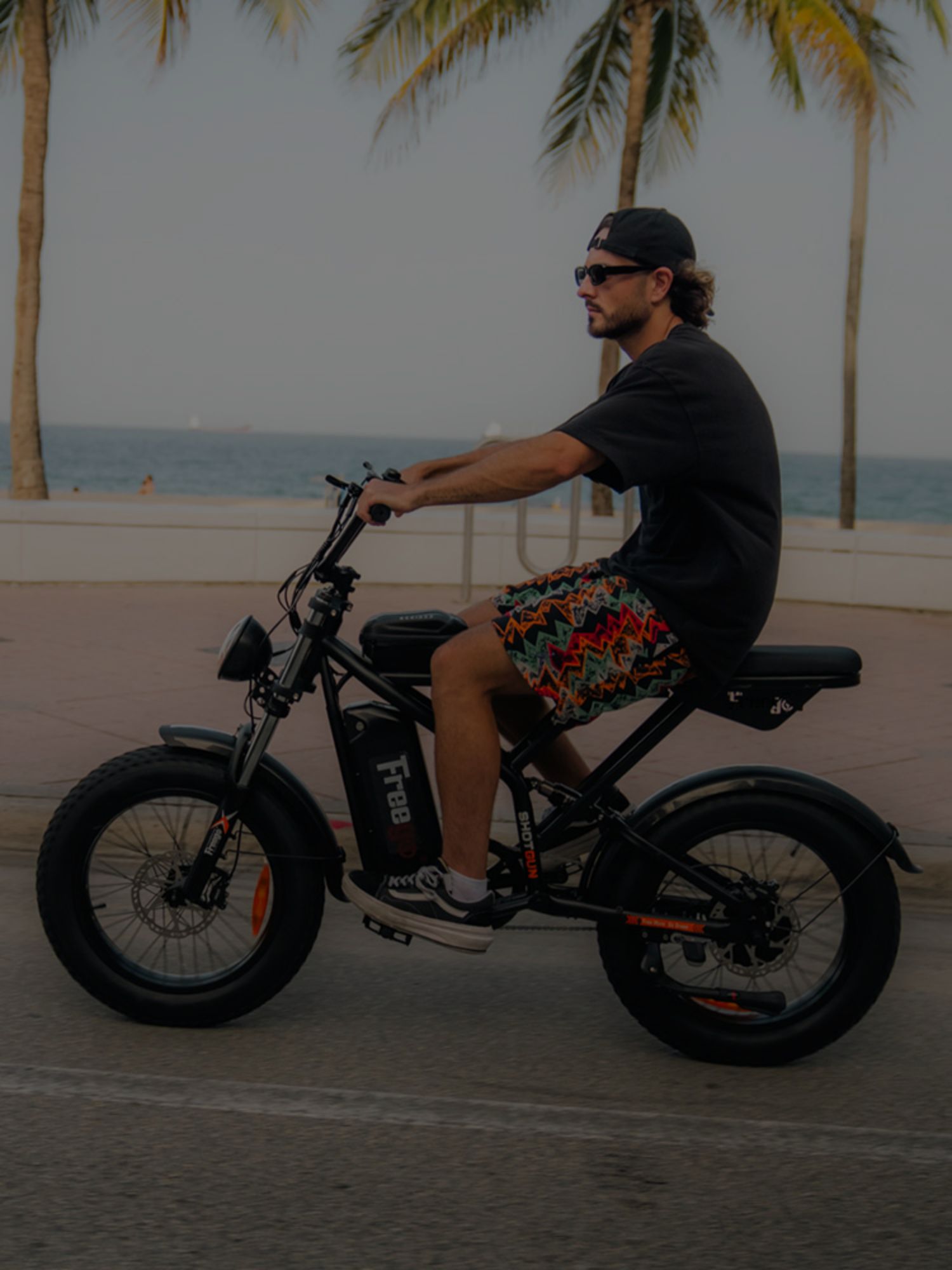 A man wearing floral pants and sunglasses with a cap riding a F2 Pro electric bike on a coastal road, palm trees in the background.